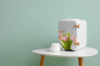 Small cosmetic refrigerator with floral decor and jars of cream on table in room