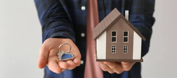 Real estate agent holding house model and key on light background, closeup