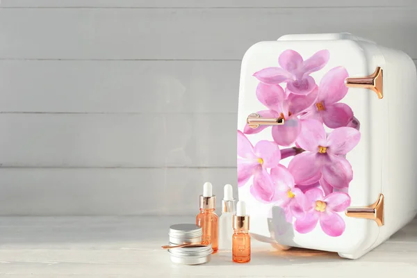 Small refrigerator with floral print and cosmetic products on light wooden background