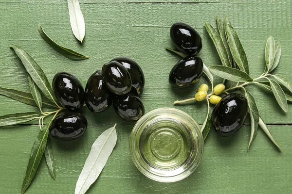 Composition with tasty black olives, bowl of oil and leaves on color wooden background