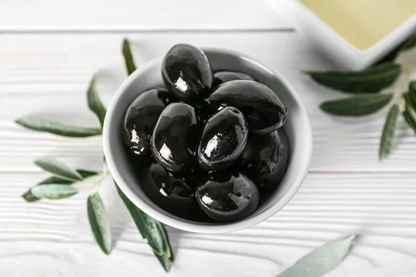 Bowl of tasty black olives and leaves on light wooden background