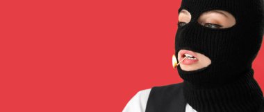 Portrait of young woman in balaclava and with burning match in mouth on red background with space for text