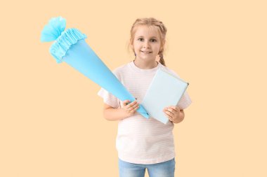 Cute little girl with blue school cone and book on beige background