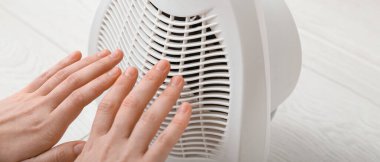 Woman warming hands near electric fan heater, closeup