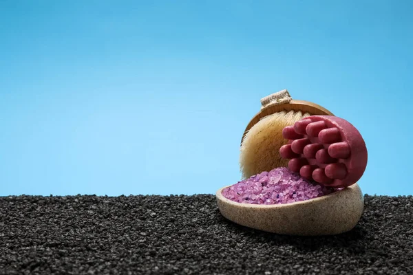 Bowl of sea salt, massage brush and soap on black sand against blue background