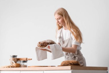 Young woman with fresh bread and napkin at table on light background
