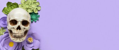 Human skull and handmade paper flowers on lilac background with space for text