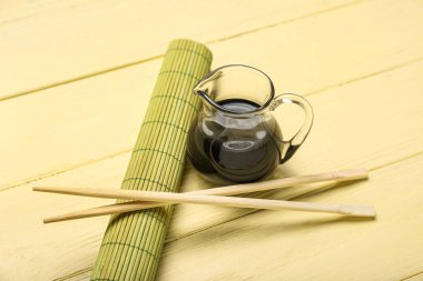 Jug of soy sauce, chopsticks and bamboo mat on yellow wooden background