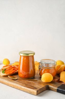 Wooden board with jars of tasty apricot jam on light background