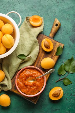 Wooden board with bowl of tasty apricot jam and fruits on green background