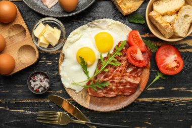 Plate with tasty fried eggs, bacon, tomatoes and ingredients on dark wooden background