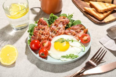 Plate with tasty fried egg, bacon, tomatoes and greens on table