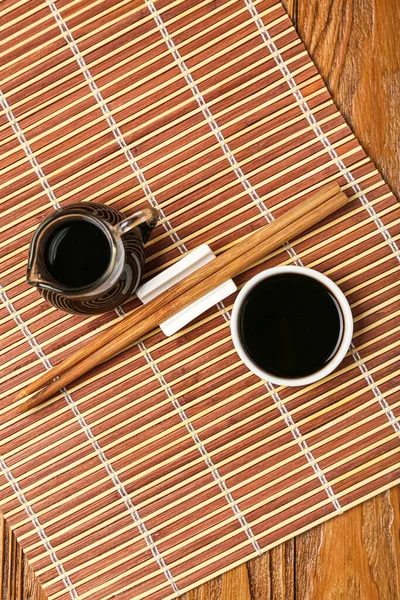 Jug and bowl of soy sauce, chopsticks and bamboo mat on wooden background