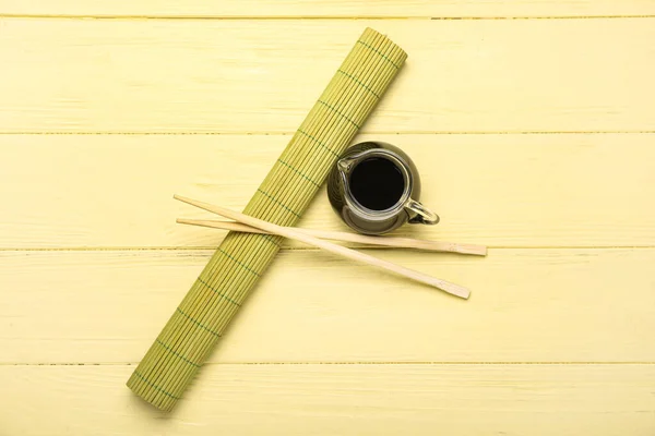 Jug of soy sauce, chopsticks and bamboo mat on yellow wooden background