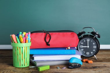 Red pencil case with school stationery and alarm clock on table near chalkboard