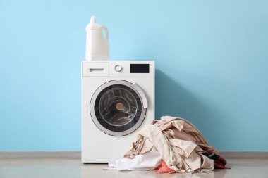 Stack of clothes and washing machine with detergent near blue wall
