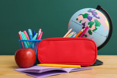 Red pencil case with school stationery and apple on table near green chalkboard