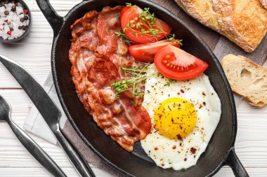 Frying pan with tasty fried egg, bacon and tomatoes on light wooden background, closeup