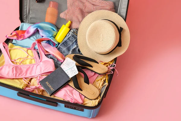 Open suitcase with clothes, passport, tickets and beach accessories on pink background
