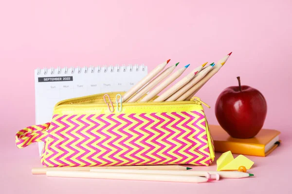 Case with pencils, paper plane, apple and calendar on pink background