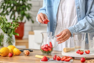 Woman putting strawberry into jug for tasty lemonade in kitchen