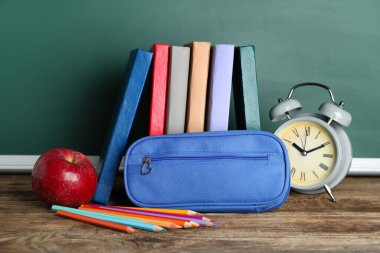 Pencil case with alarm clock, books and apple on table near chalkboard