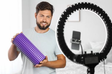 Young man with foam roller recording video in bedroom