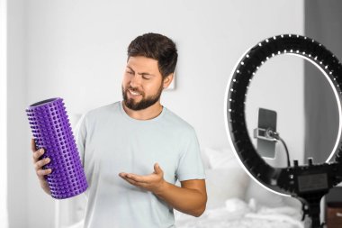 Young man with foam roller recording video in bedroom