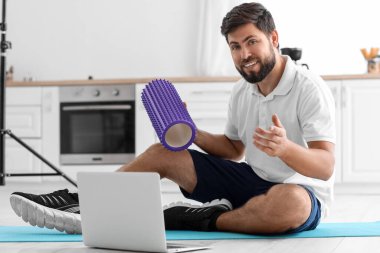 Young man with foam roller and laptop in kitchen