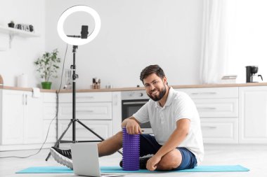 Young man with foam roller and laptop in kitchen