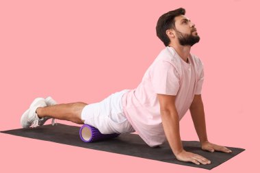 Young man training with foam roller on pink background