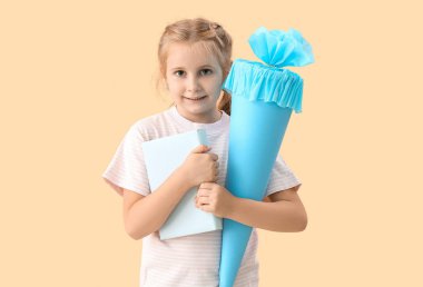 Cute little girl with blue school cone and book on beige background
