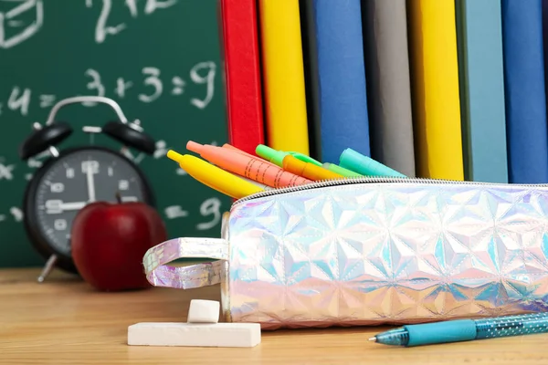 Pencil case with school stationery, alarm clock and apple on table near green chalkboard