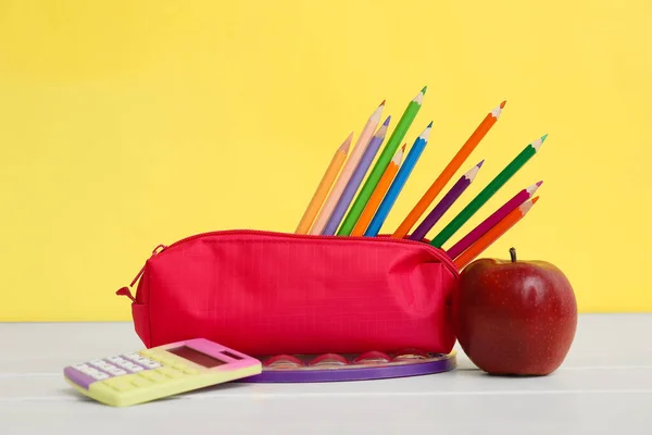 Red pencil case with school stationery and apple on table against yellow background