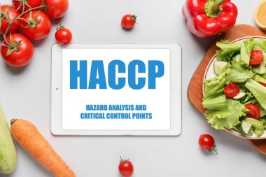 Tablet computer with text HAZARD ANALYSIS AND CRITICAL POINTS and fresh vegetables on light background