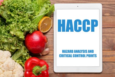 Tablet computer with text HAZARD ANALYSIS AND CRITICAL POINTS and fresh vegetables on wooden background