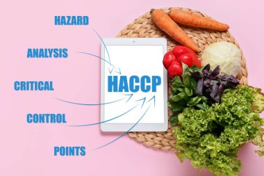 Tablet computer with text HAZARD ANALYSIS AND CRITICAL POINTS and fresh vegetables on pink background