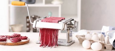 Pasta maker with red dough and ingredients on kitchen table