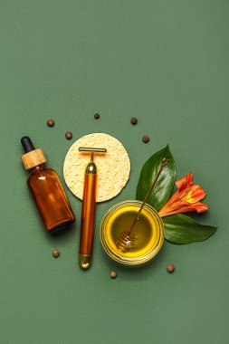 Facial massage tool with cosmetic dropper bottle, honey, sponge and flower on green background