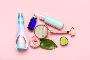Facial massage tool with cosmetic products, cucumber slice and flower on pink background
