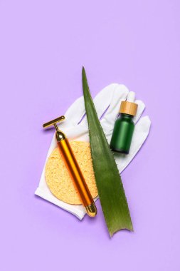 Facial massage tool with cosmetic bottle, aloe vera leaf, sponge and glove on lilac background
