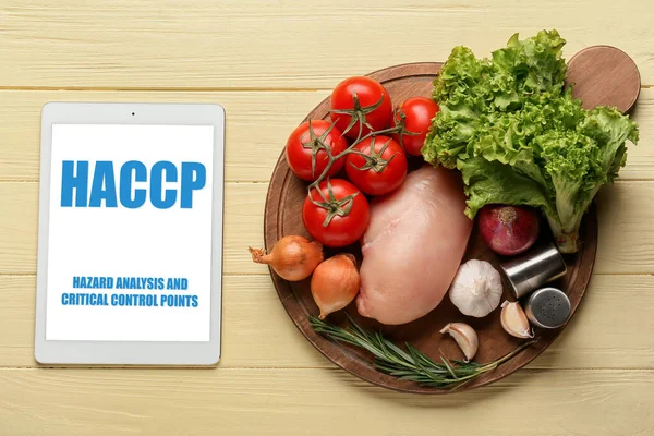 Tablet computer with text HAZARD ANALYSIS AND CRITICAL POINTS and fresh products on yellow wooden background