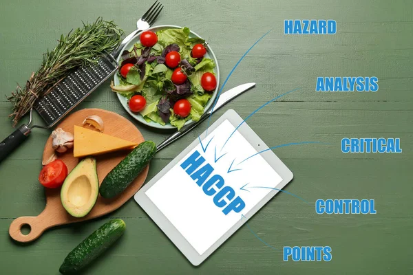 Tablet computer with text HAZARD ANALYSIS AND CRITICAL POINTS and fresh products on green wooden background