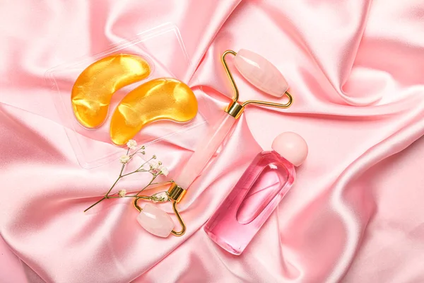 Facial massage tool with cosmetic bottle, under-eye patches and flowers on pink fabric background