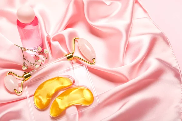 Facial massage tool with cosmetic bottle, under-eye patches and flowers on pink fabric background