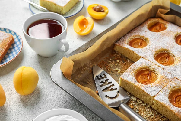 Baking tray with tasty apricot pie and cup of tea on light table, closeup