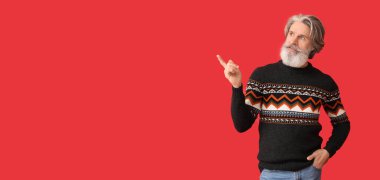 Handsome senior man in knitted sweater showing something on red background with space for text