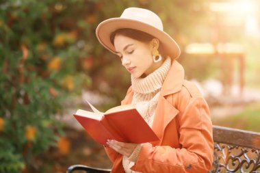 Beautiful young woman reading interesting book in autumn park