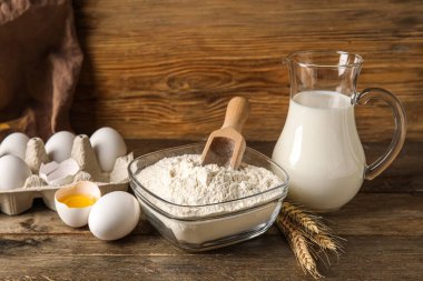 Bowl with flour, eggs and jug with milk on table against wooden background