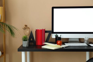 Modern workplace with computer, bookend, apple and pen cup near beige wall in room